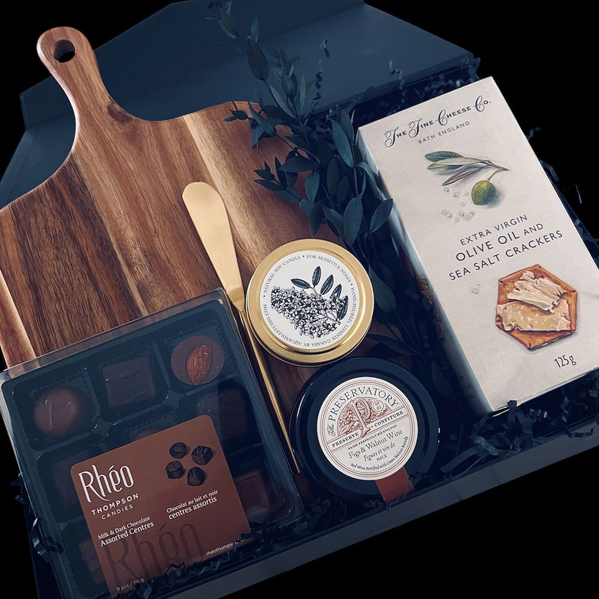 A gourmet gift collection featuring a wooden cutting board, a gold spreader knife, a box of Rheo Thompson Chocolates, Extra Virgin Olive Oil and Sea Salt Crackers, Preservatory Fig and Walnut Wine Charcuterie Spread and a Muse Scented Candle arranged in an elegant black magnetic box.