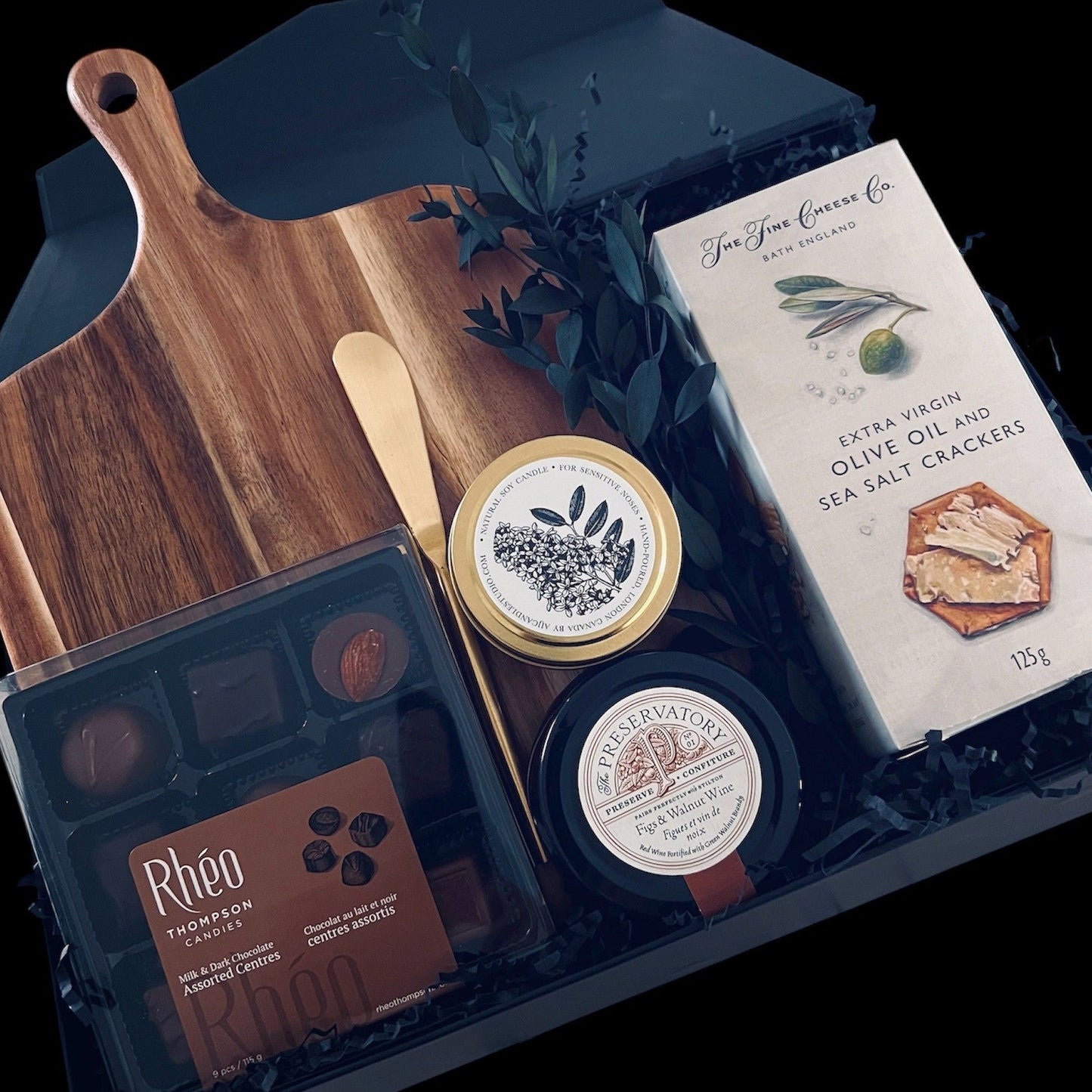 A gourmet gift collection featuring a wooden cutting board, a gold spreader knife, a box of Rheo Thompson Chocolates, Extra Virgin Olive Oil and Sea Salt Crackers, Preservatory Fig and Walnut Wine Charcuterie Spread and a Muse Scented Candle arranged in an elegant black magnetic box.